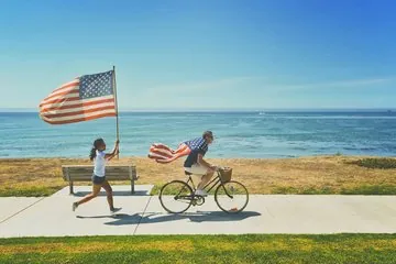A man rides a bicycle near the ocean with an American flag draped over his shoulders while a girl runs beside him holding a large U.S. flag.